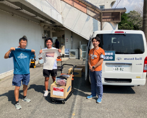 アウトドア義援隊の男性と、支援したTシャツを広げて持つ男性二人が並んでいます。