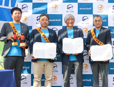 青森県鰺ヶ沢町、深浦町と株式会社モンベルの締結式の様子。
左から立会人の秋田県八峰町・堀内満也 町長、青森県深浦町・平沢一臣 町長、モンベル代表・辰野勇、青森県鰺ヶ沢町・平田衛 町長がクマの像、包括協定書を掲げています。